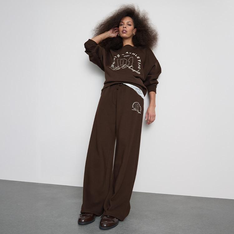 Person wearing brown matching sweatshirt and wide-leg trousers with logo detailing against grey background.