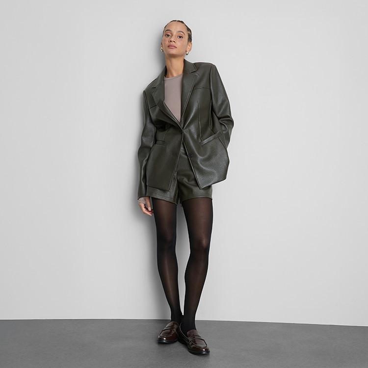 Person wearing dark green leather blazer and shorts with black tights and brown loafers.