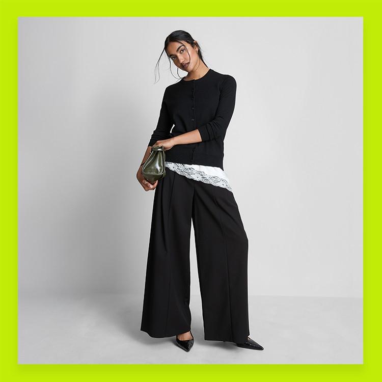 Person wearing black cardigan with lace trim detail, wide-leg trousers and holding a green handbag.