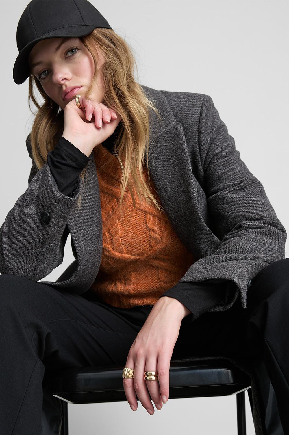 Person wearing grey blazer, orange knitted jumper and black cap, seated with gold rings on fingers.