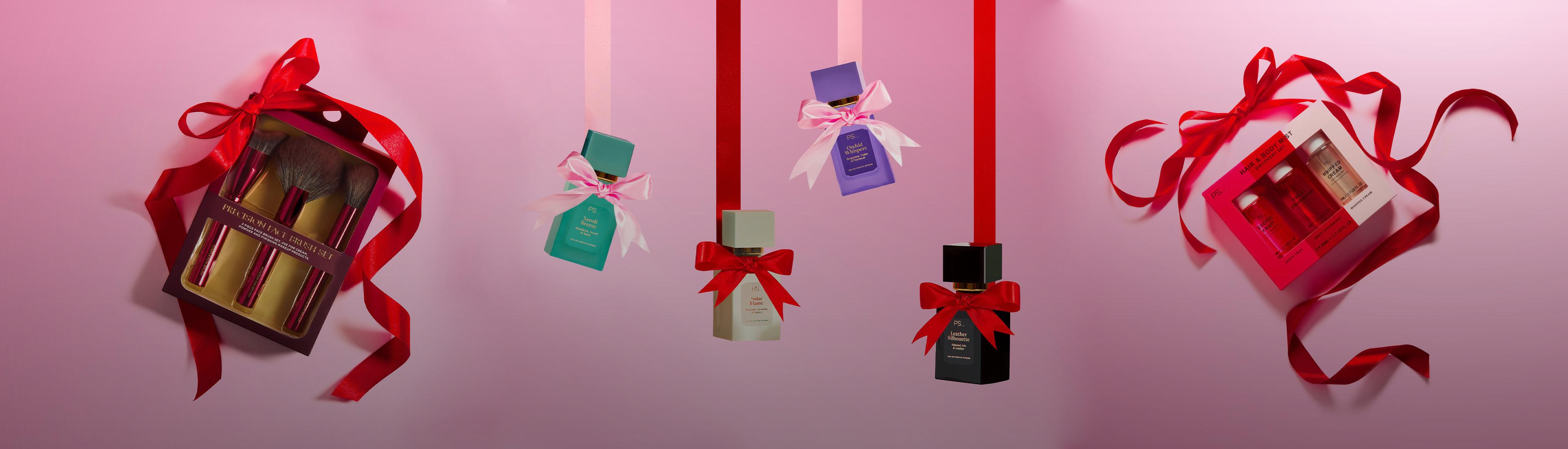 Collection de cadeaux de fêtes : pinceaux de maquillage et flacons de parfum ornés de rubans rouges et roses sur fond rose.