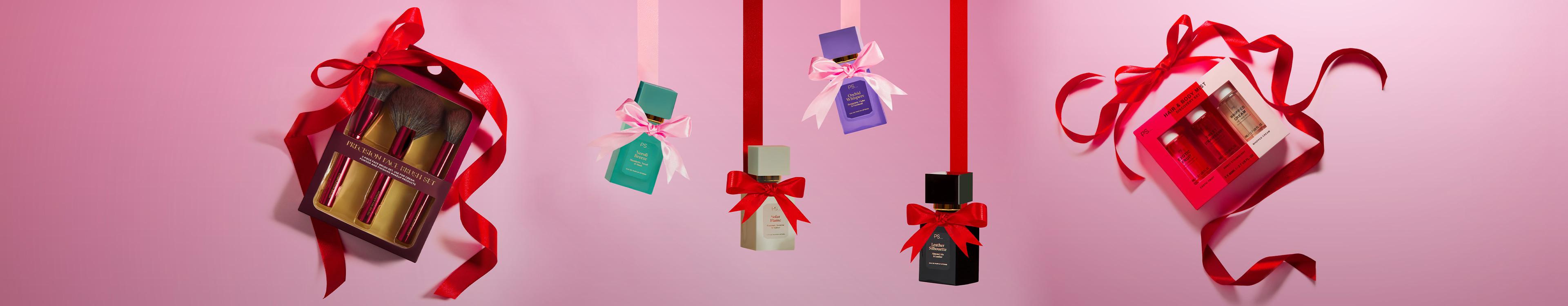 Holiday beauty gifts including makeup brushes and perfume bottles decorated with red ribbons on pink background.