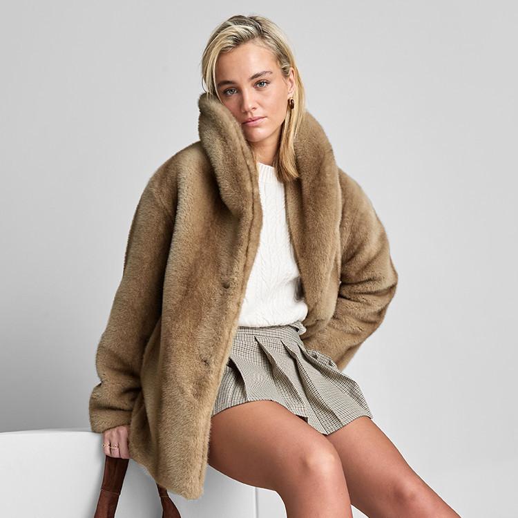 Person wearing luxurious camel faux fur coat over white jumper and checked shorts, seated against grey background.