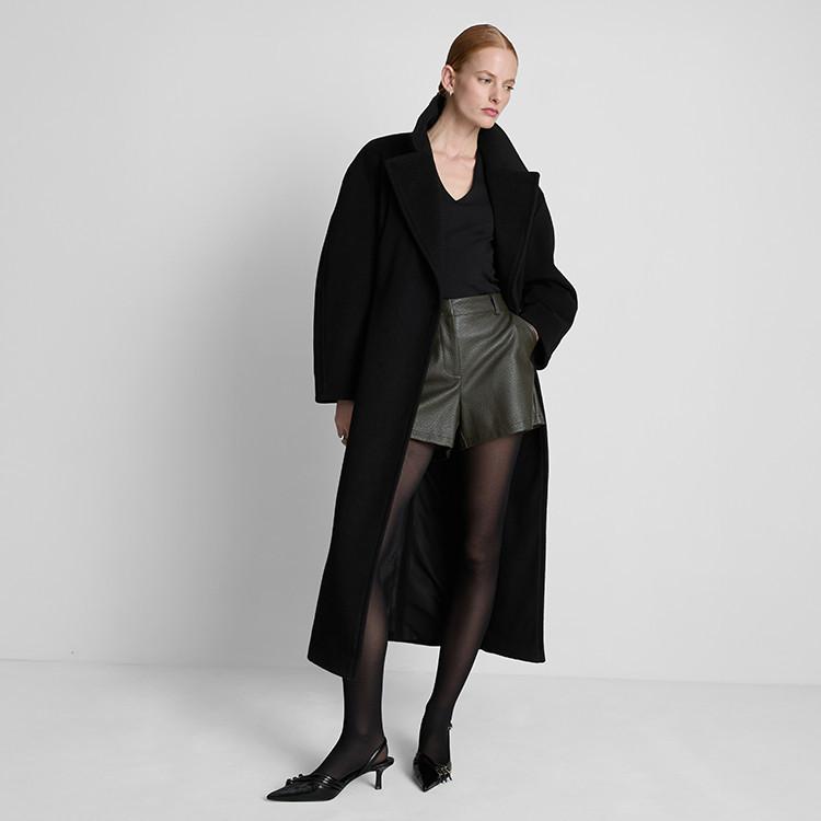 Model wearing long black wool coat with green leather shorts, black top and sheer tights.
