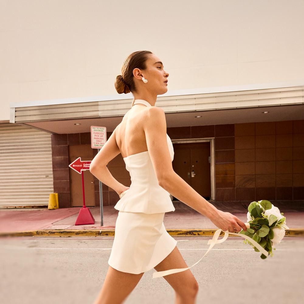 Frau läuft selbstbewusst in weißem schulterfreiem Top mit Schößchen und passendem Rock mit einem Blumenstrauß in der Hand.