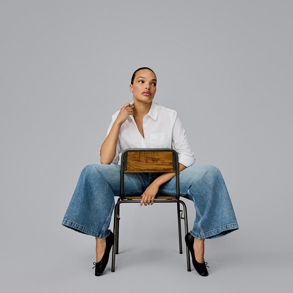 Person wearing white button-up shirt and wide leg jeans seated on a wooden chair against grey backdrop.