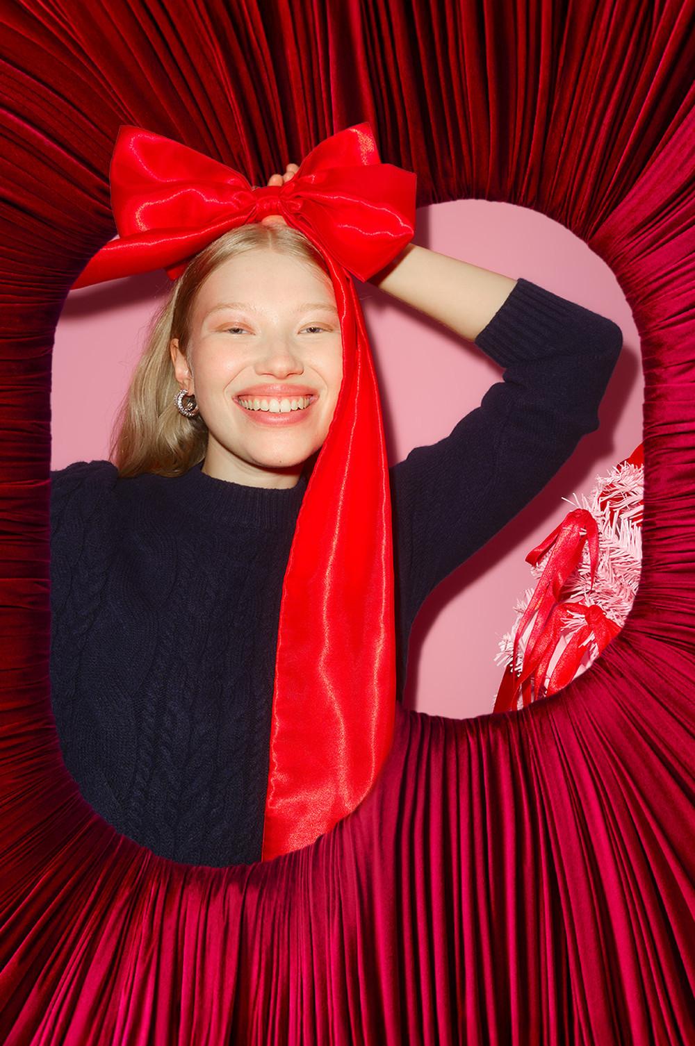 Personne portant un pull bleu marine avec un grand nœud rouge et entourée de draperies rouges vives sur fond rose.