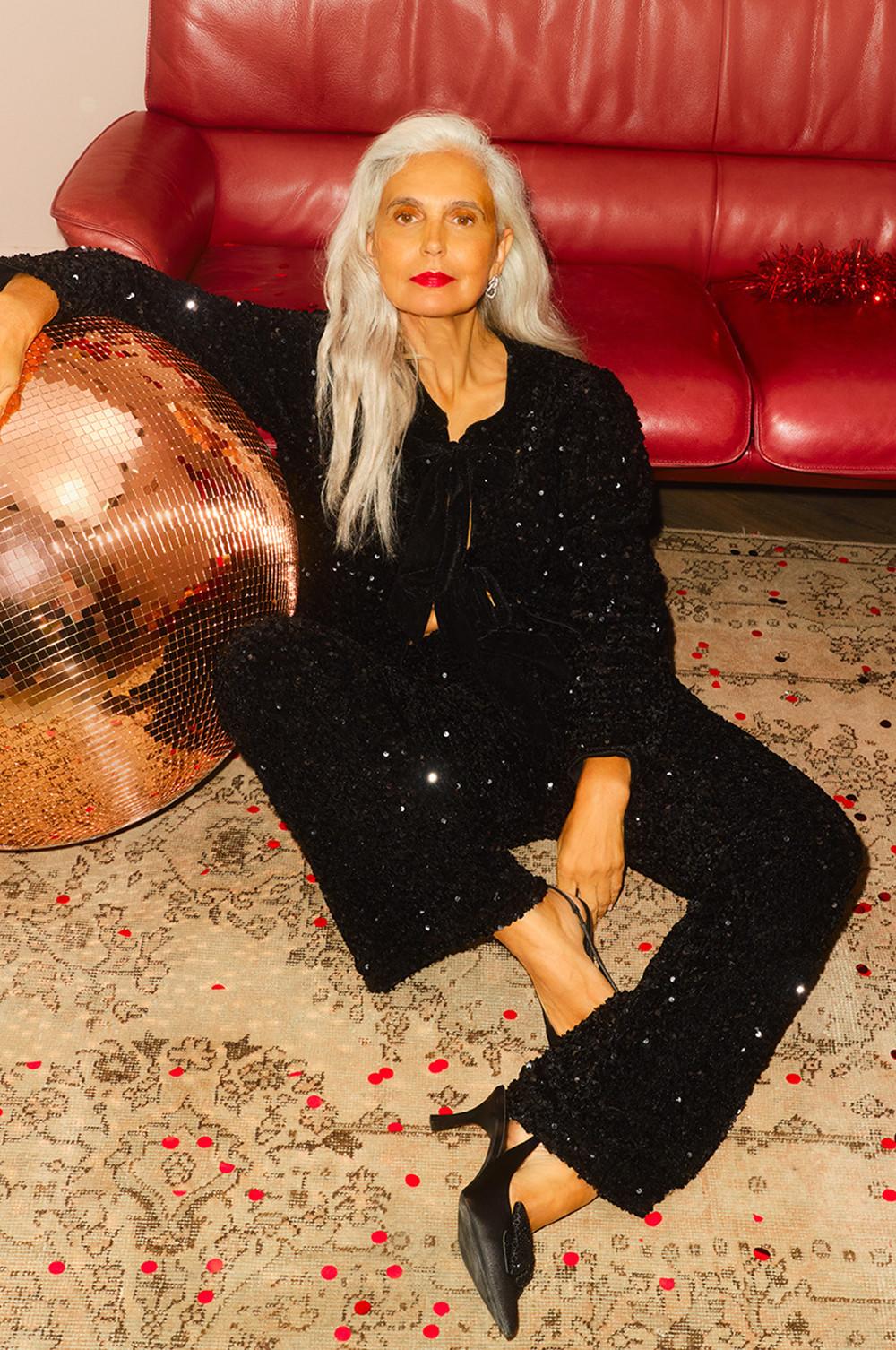 Personne portant une tenue noire scintillante, assise à côté d’une boule à facettes cuivrée sur un canapé en cuir rouge.