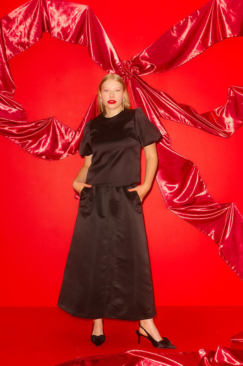Élégant ensemble deux pièces en satin noir, composé d’un haut à manches bouffantes et d’une jupe longue, sur un fond spectaculaire en tissu rouge.
