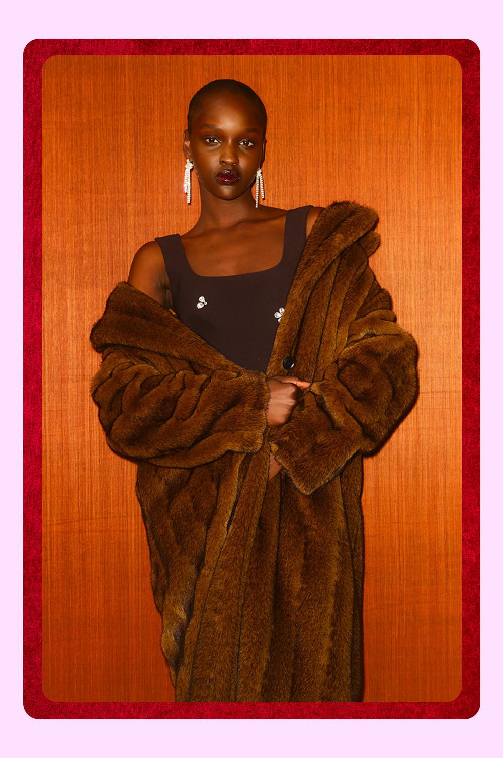 Mannequin portant un manteau en fausse fourrure marron sur une robe foncée ornée de strass, avec des boucles d’oreilles pendantes.