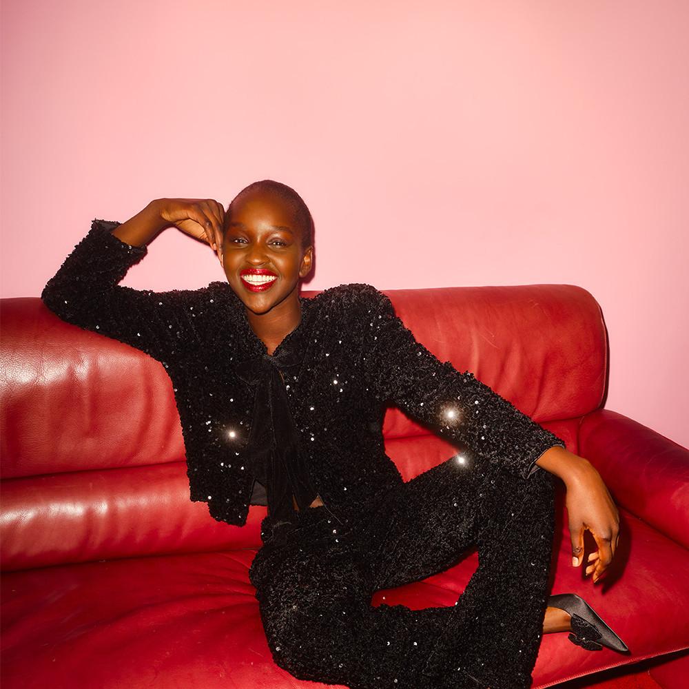 Personne portant une tenue noire scintillante et assise sur un canapé en cuir rouge sur fond rose.