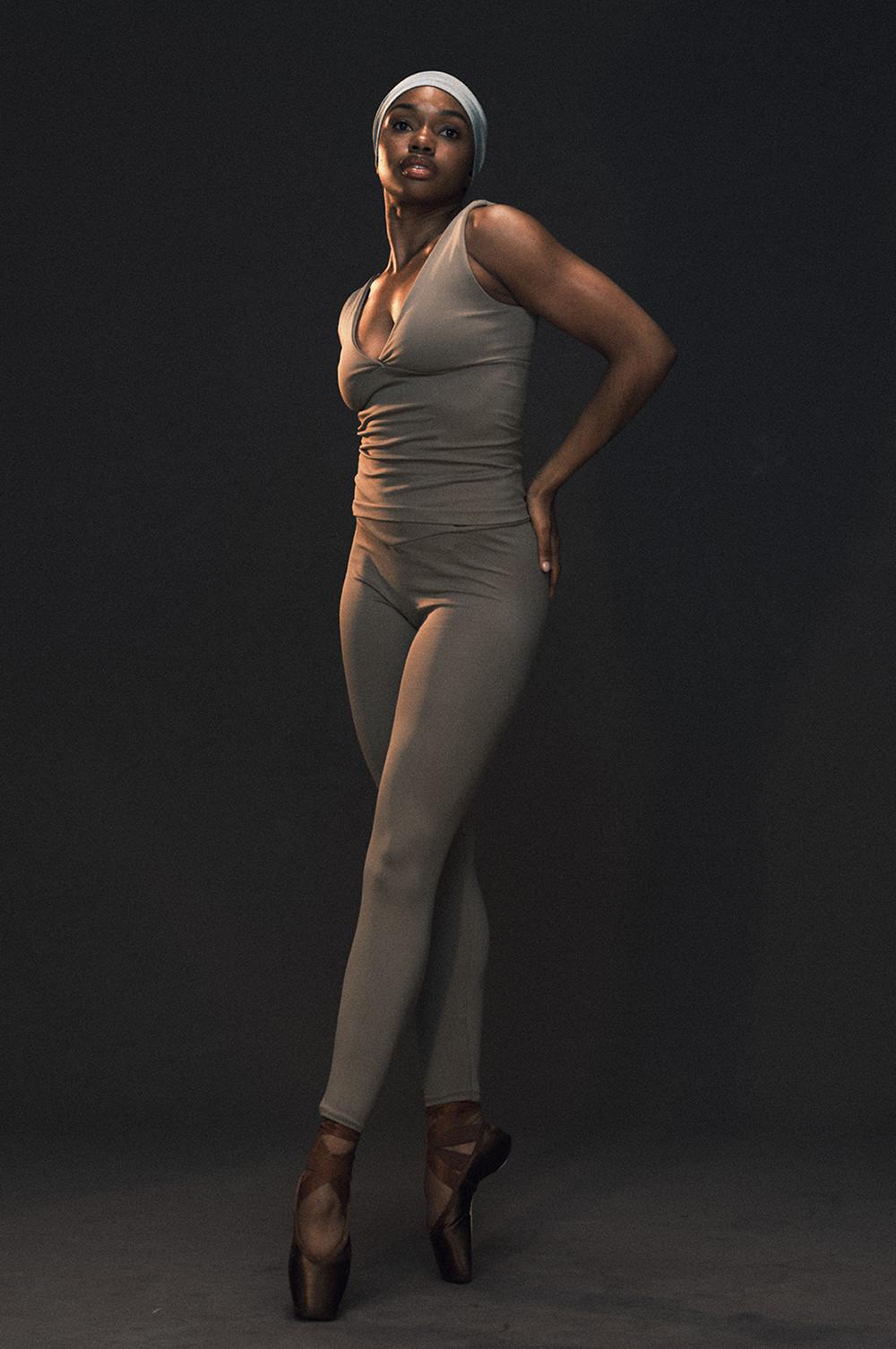 Ballet dancer in beige leotard and tights with white headwrap, posing en pointe against dark backdrop.