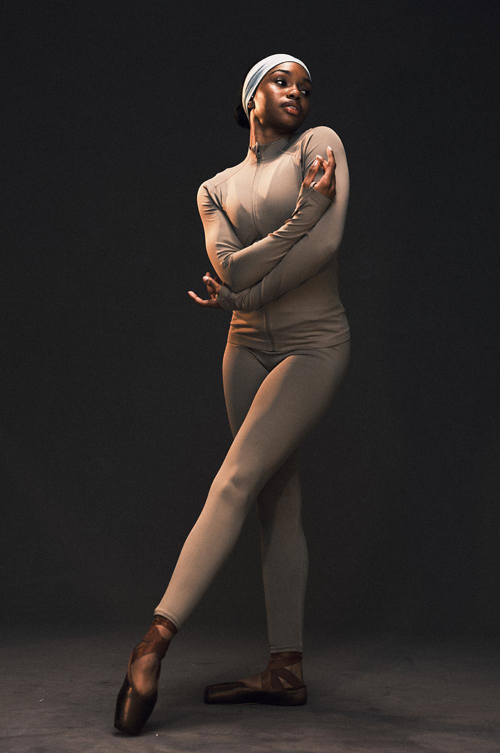Dancer in beige fitted bodysuit and white head covering performing ballet pose on pointe shoes.