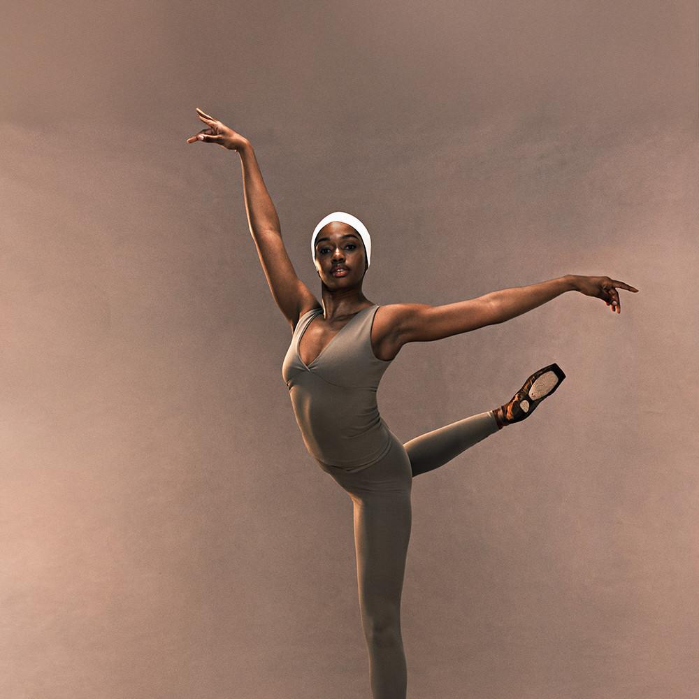 Dancer in taupe leotard and white headwrap performing an arabesque position with arms extended.