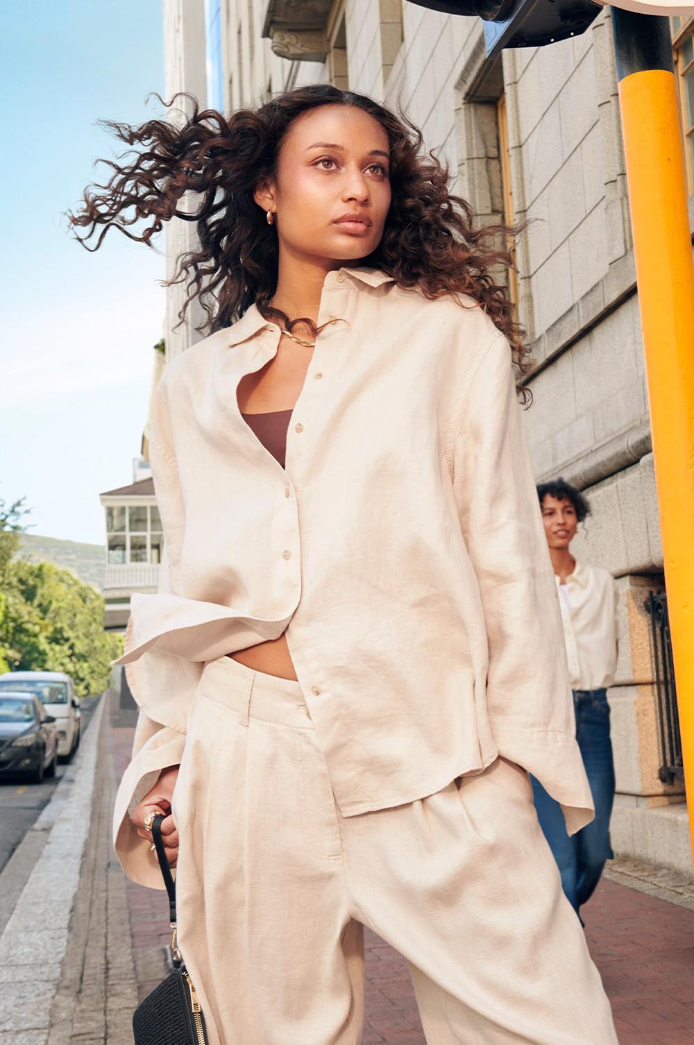Modelo com camisa de linho e calças a condizer em cor creme, a caminhar numa rua da cidade, com o cabelo encaracolado ao vento.