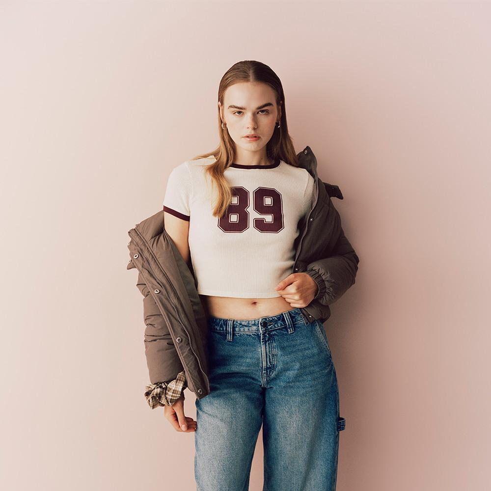 Modella che indossa una maglietta corta bianca con il numero "89" al centro, blue jeans e giacca.