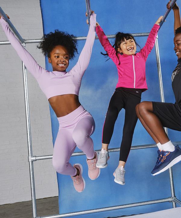 Crianças com a gama da roupa de desporto Da esquerda para a direita: conjunto de leggings e top lilás, leggings pretas e casaco cor-de-rosa, conjunto de calções pretos e top.