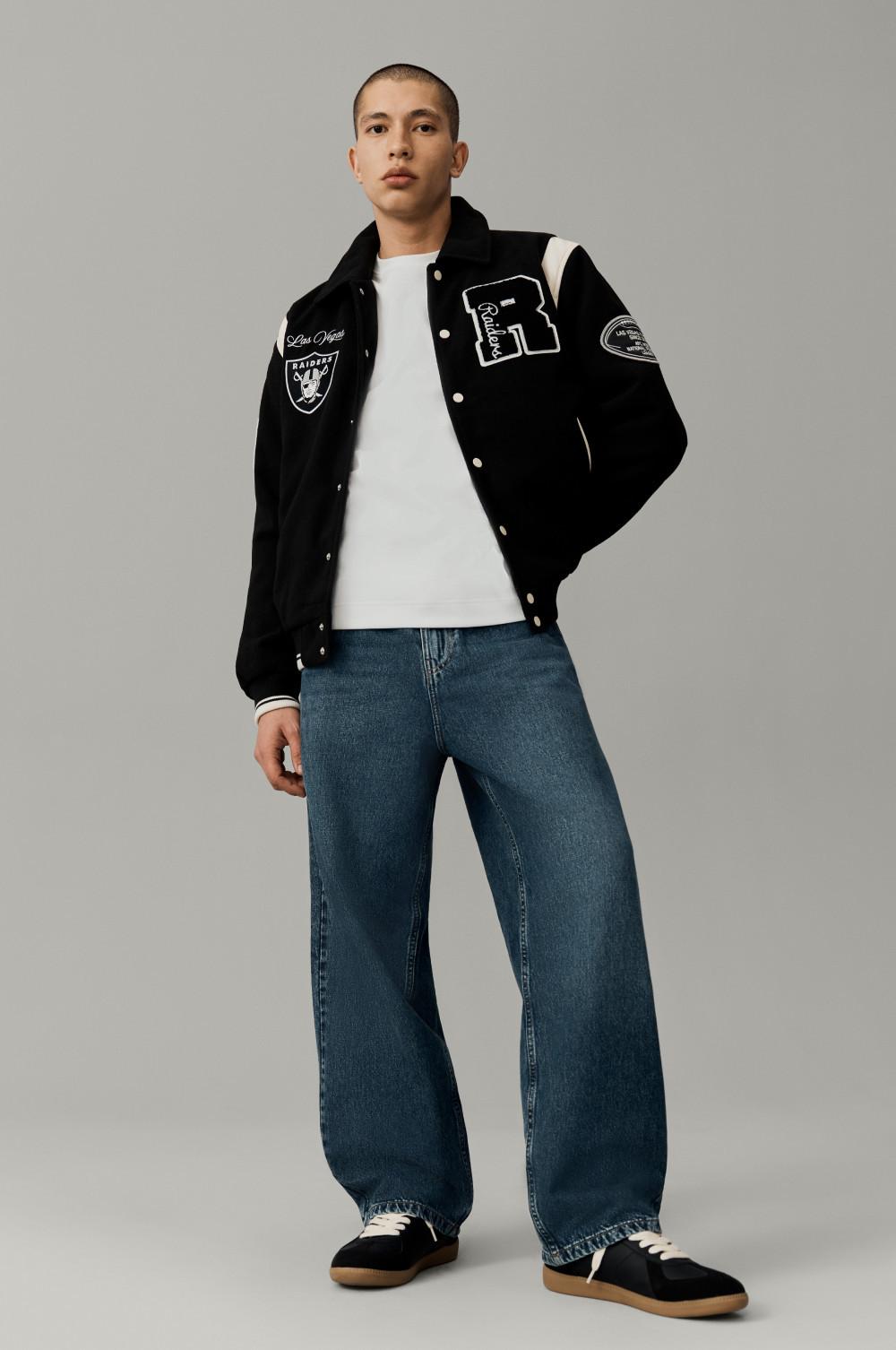 Person wearing black varsity jacket with Raiders logo, white T-shirt and wide-leg blue jeans.