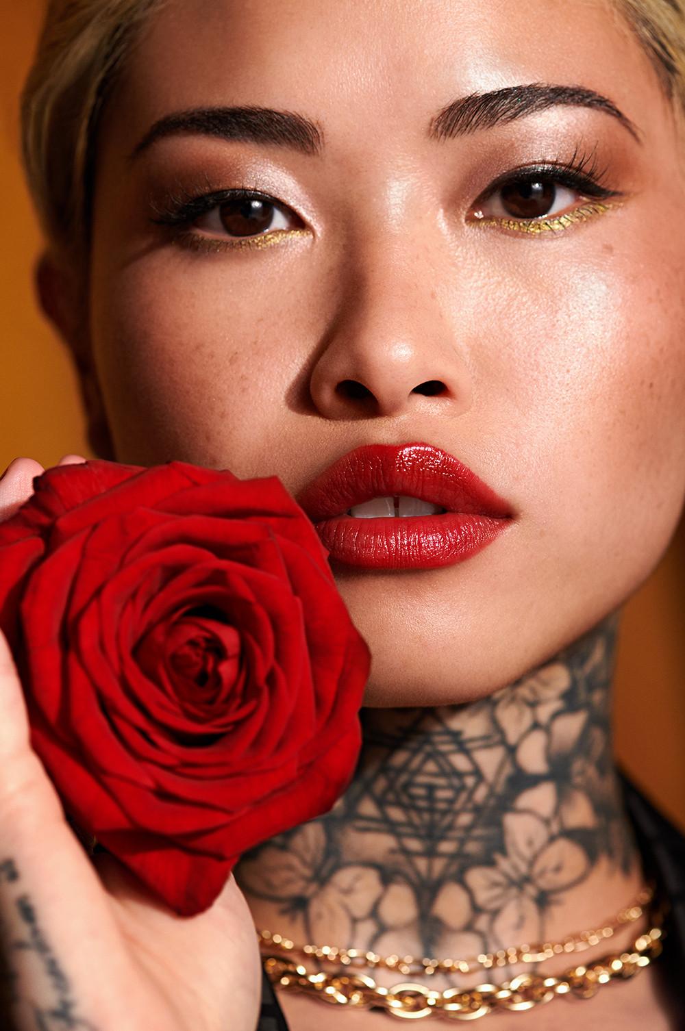 Model holds red rose, whilst wearing red lipstick, gold and black eyeliner and a gold chain necklace