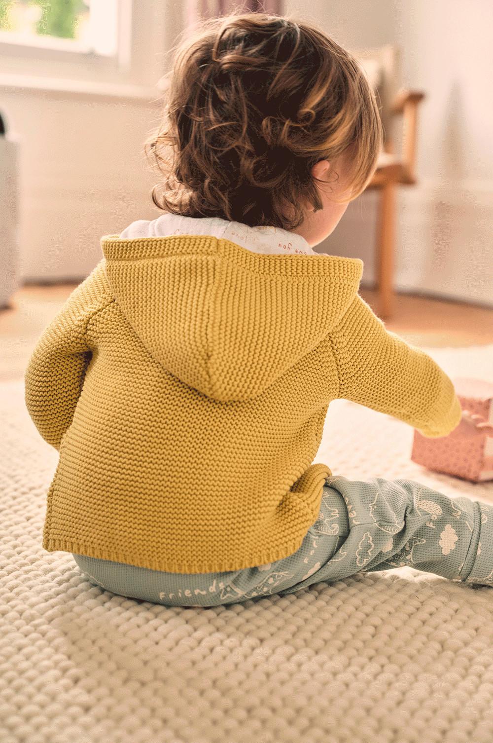 Sitzendes Baby in senfgelbem Cardigan und blauer Hose