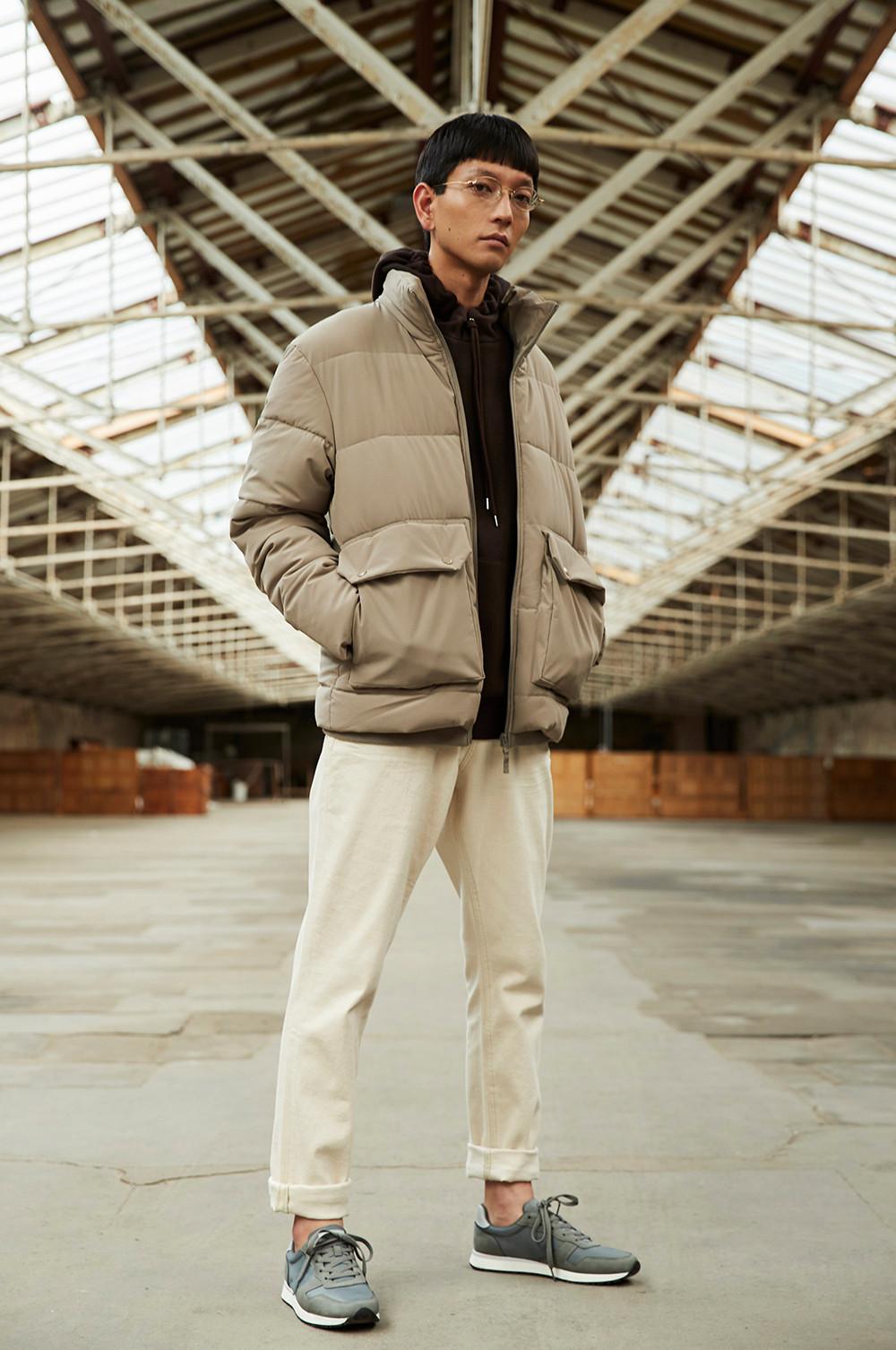 Model stands in an empty warehouse, wearing cream joggers, black hoodie and beanie and a stone colou