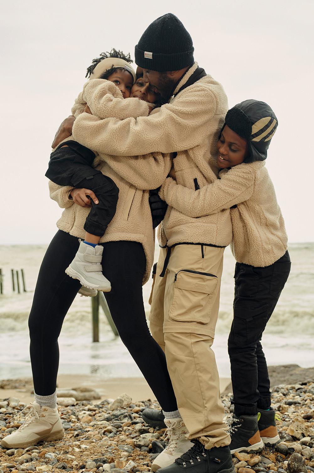 Familia en la playa con chaquetas polares en cámel, pantalones térmicos y cargo, y gorros negros.