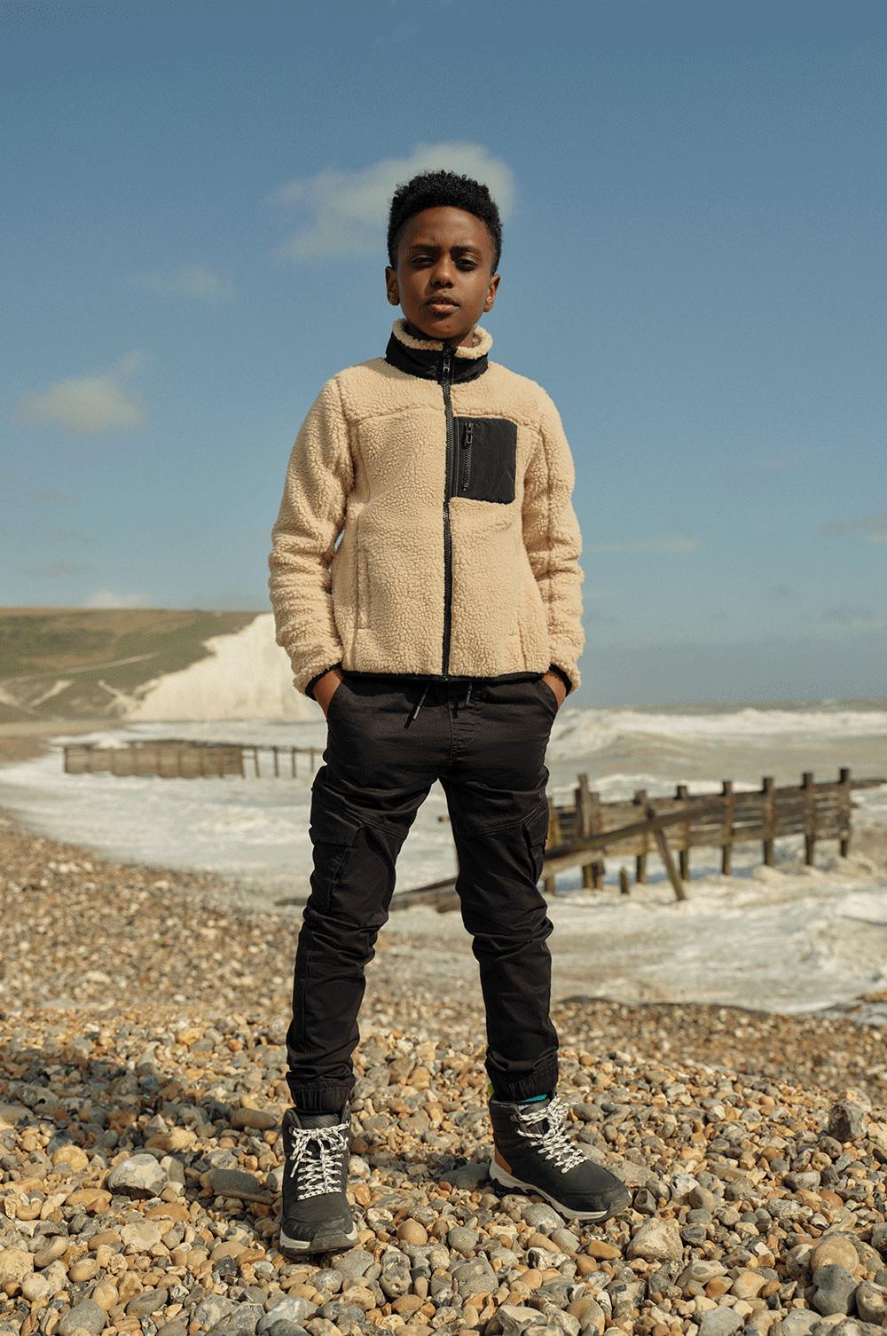 Fant stoji na plaži, oblečen v termo hlače in mucast flis