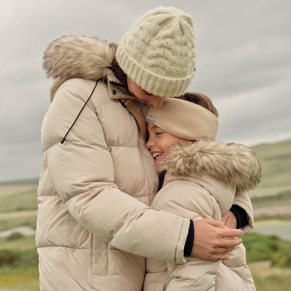 Mother and daughter hugging outside, both wear cream puffer coats, beanie hats and ear warmers