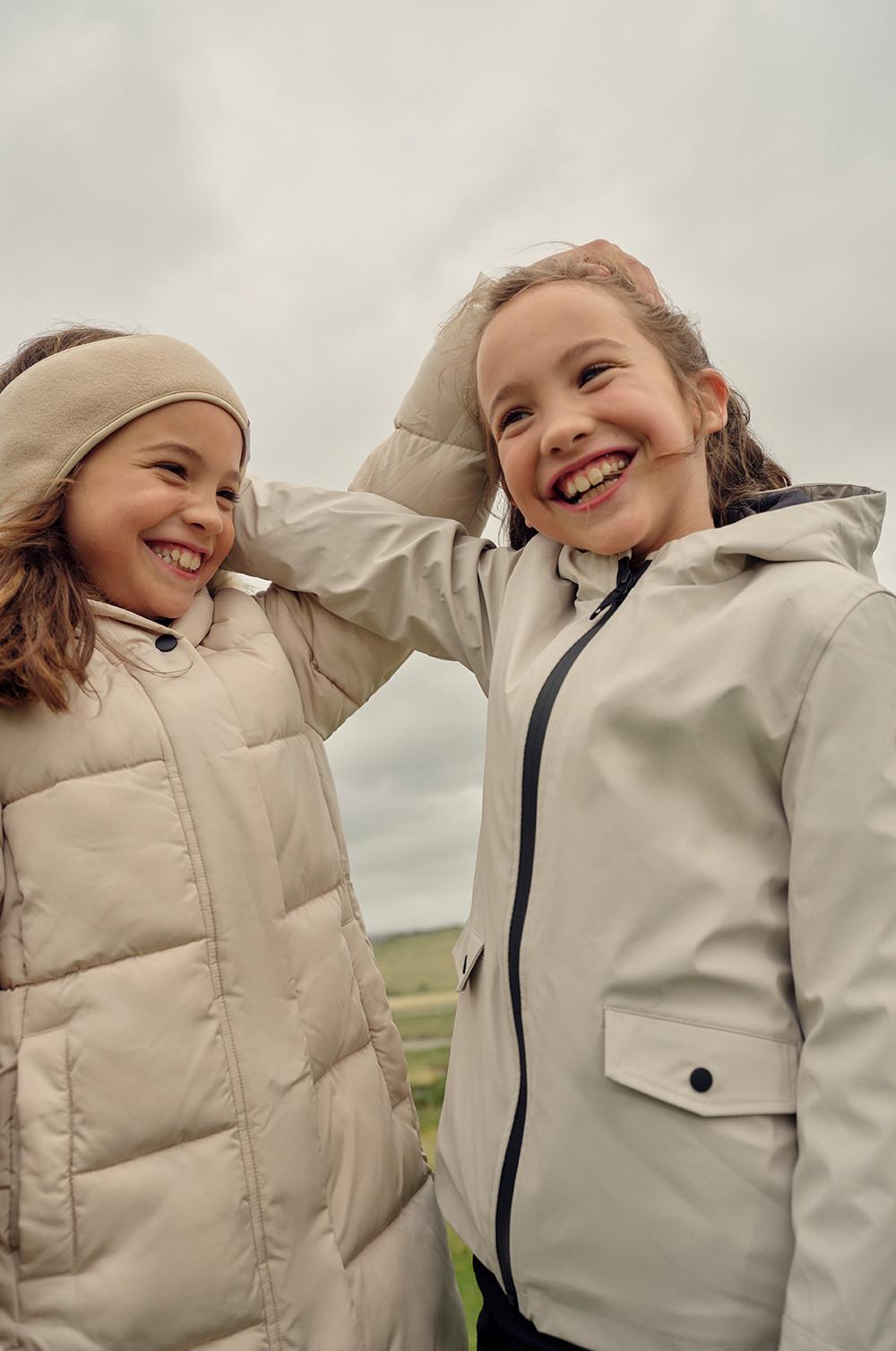 two girls wear cream puffer and rain coats while smiling