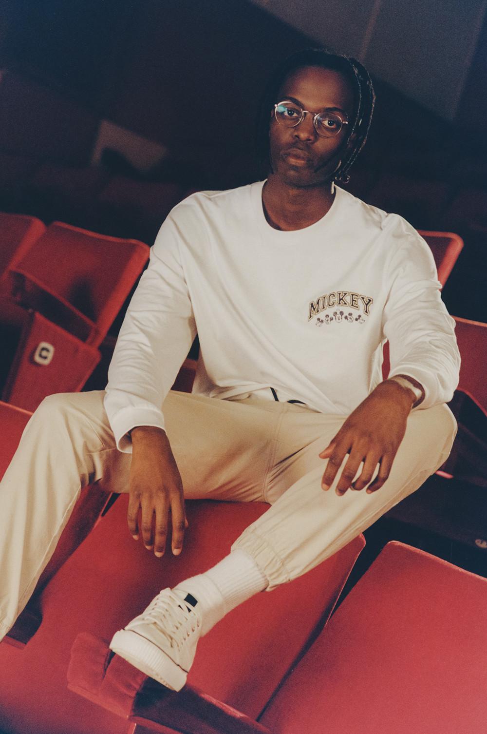 Model sits wearing a white long sleeved Mickey t-shirt, cargo joggers, white socks and trainers