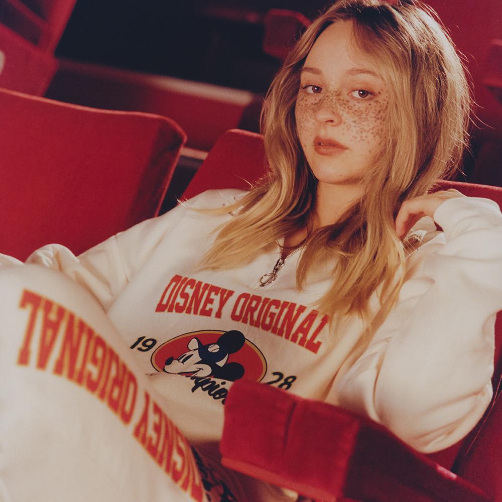 Model sits, wearing Mickey mouse white tracksuit, with a sweatshirt and cuffed joggers