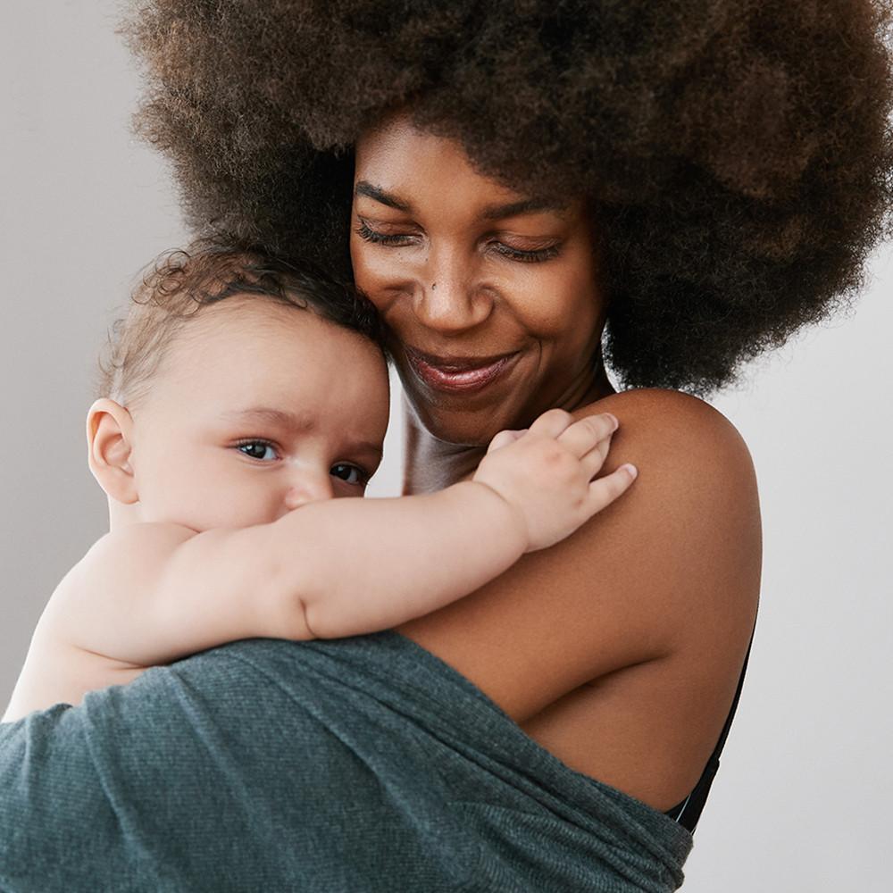 Madre sonriendo y abrazando a su bebé. Lleva una rebeca gris cayéndole de los hombros.