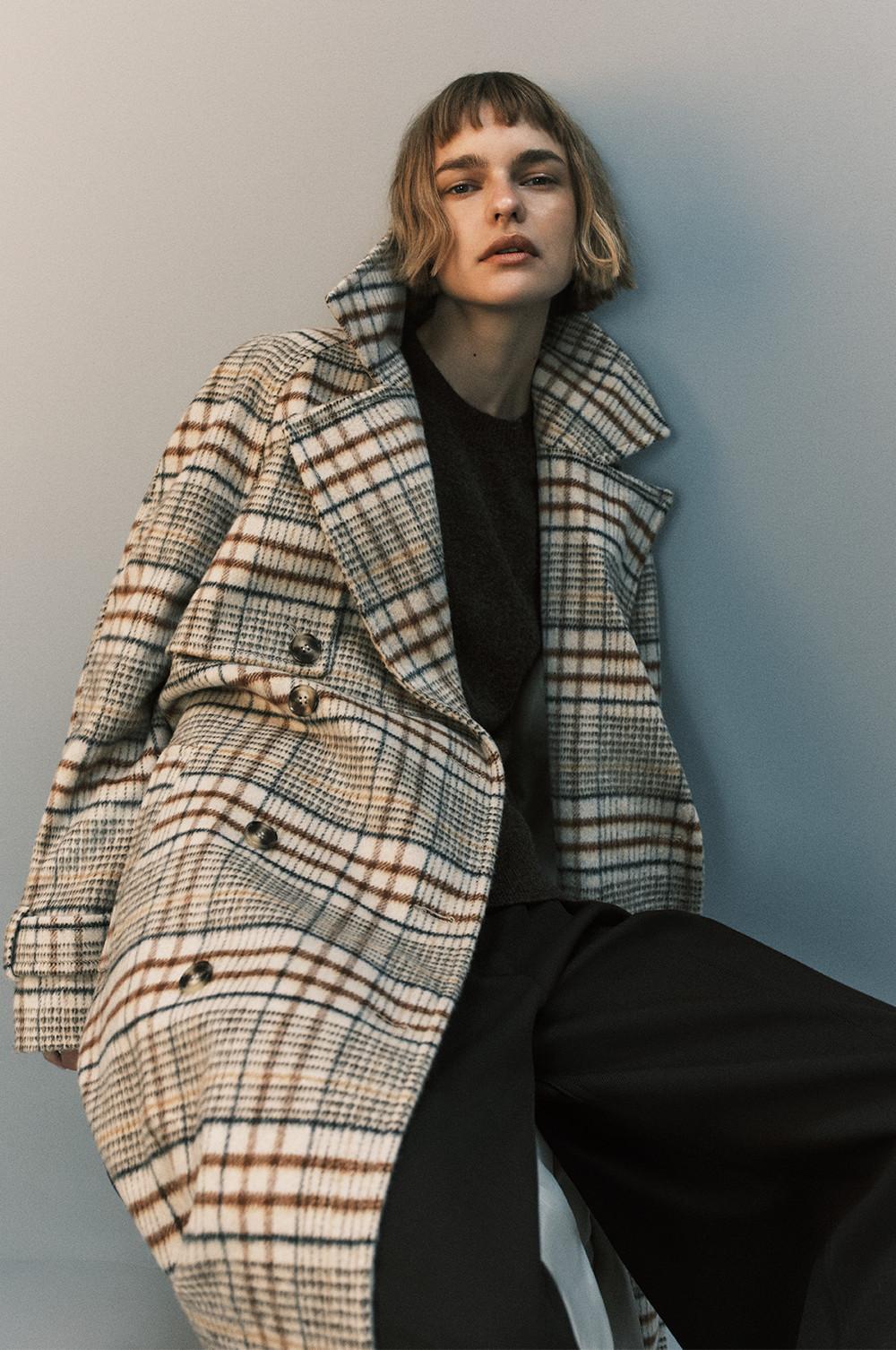 Person wearing a cream and brown checked wool coat over dark clothing against a neutral background.