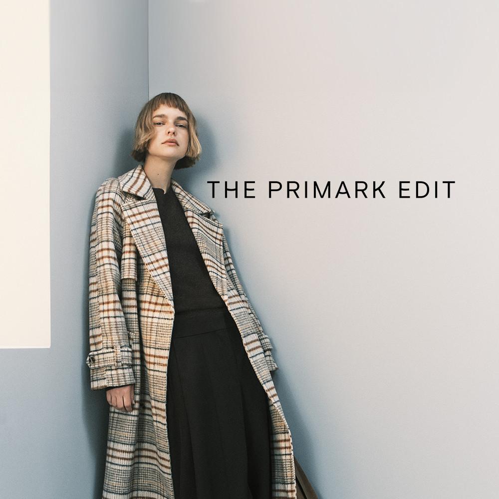 Model wearing a plaid patterned coat over a black outfit, standing against a light wall. THE PRIMARK EDIT text displayed.