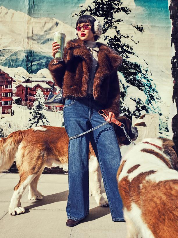 Person in brown fur jacket and wide-leg jeans holding drink while walking two dogs against winter resort backdrop.