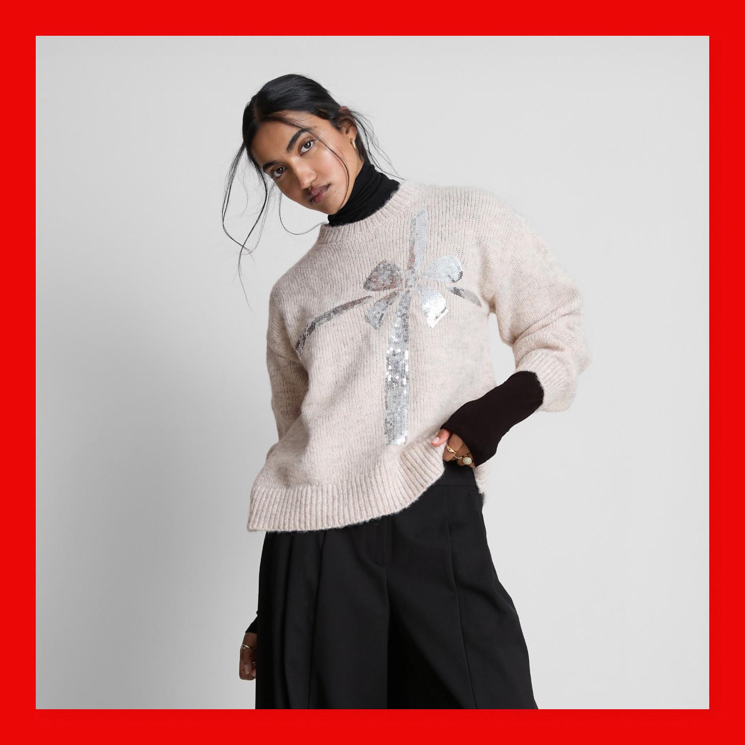 Cream knitted jumper with sequin gift bow design, worn over black turtleneck and wide-leg trousers.