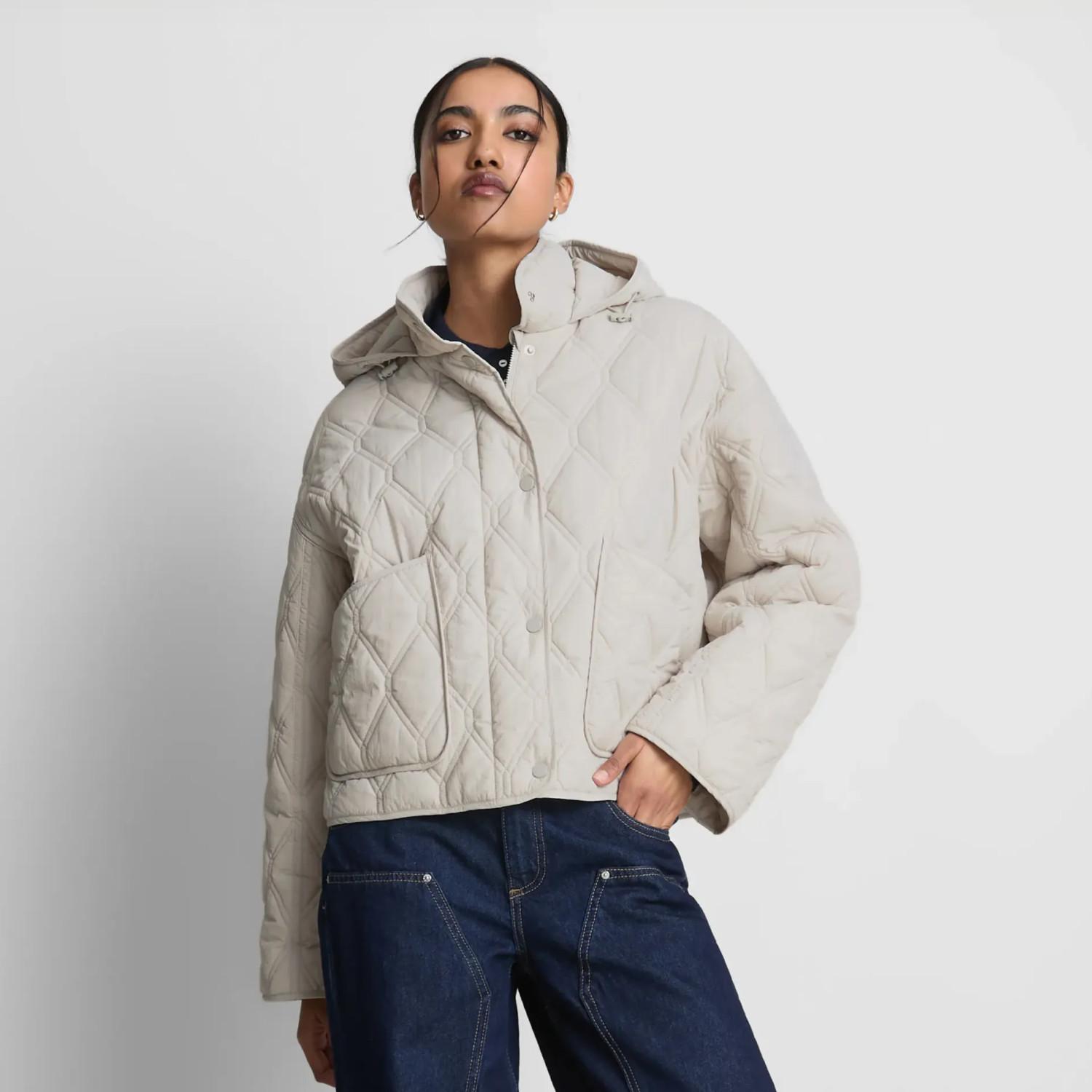 Beige quilted hooded jacket with button closure worn with dark blue jeans against a white background.