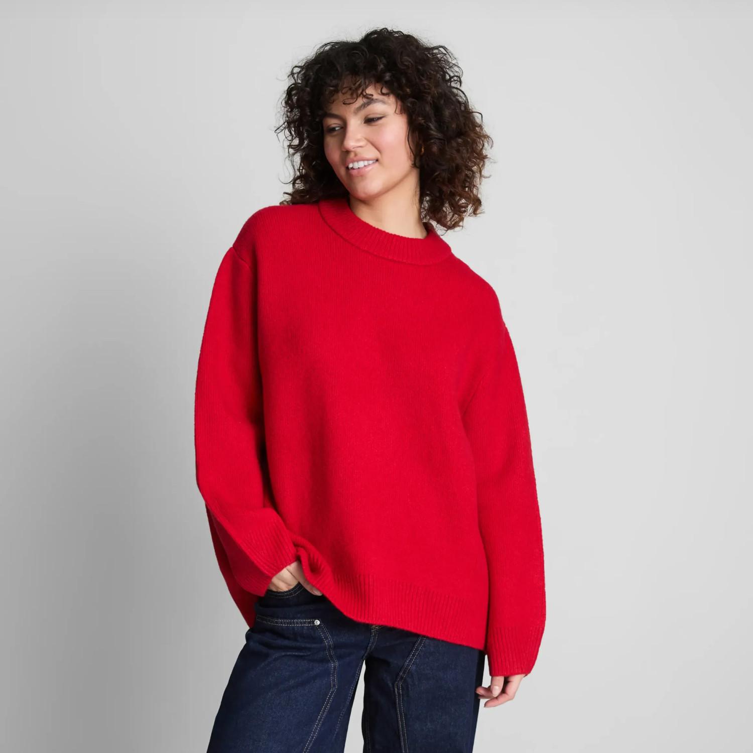 Person wearing bright red oversized jumper with crew neck and dark denim jeans against neutral background.