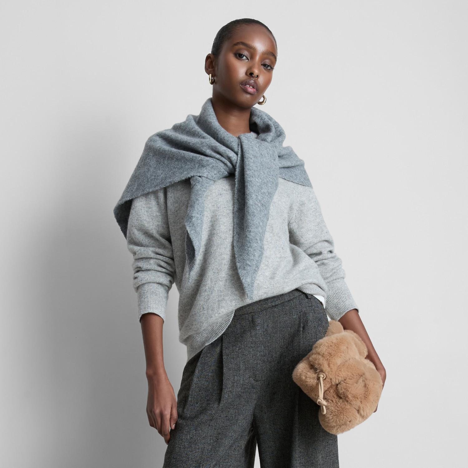 Model wearing grey knit sweater with matching scarf, dark wool trousers and holding a soft beige fur handbag.
