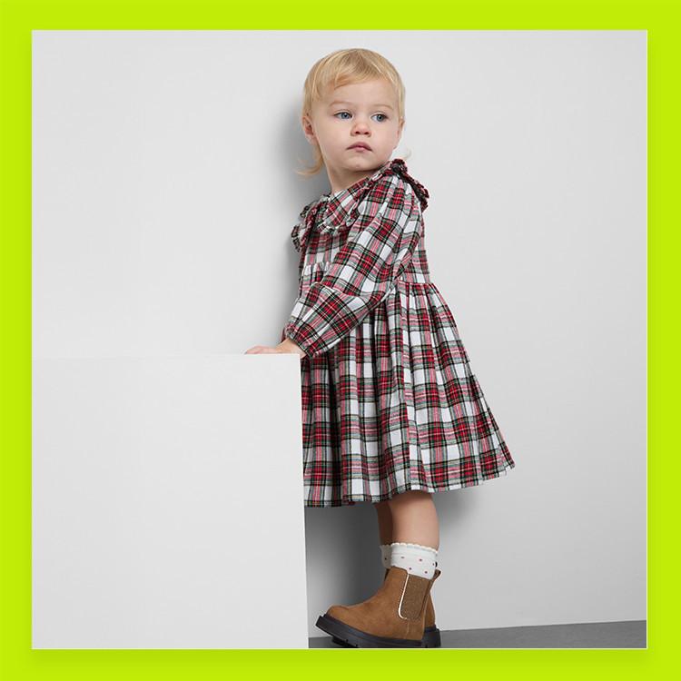 Toddler wearing red and white plaid dress with brown chelsea boots and white socks against grey background.
