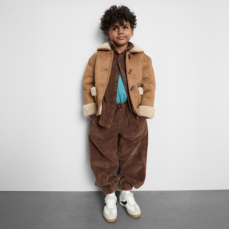 Child wearing tan shearling jacket over brown corduroy outfit with turquoise top and white trainers.