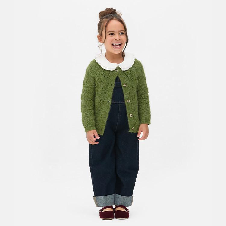 Child wearing green knitted cardigan with white collar over dark denim jumpsuit and burgundy shoes.