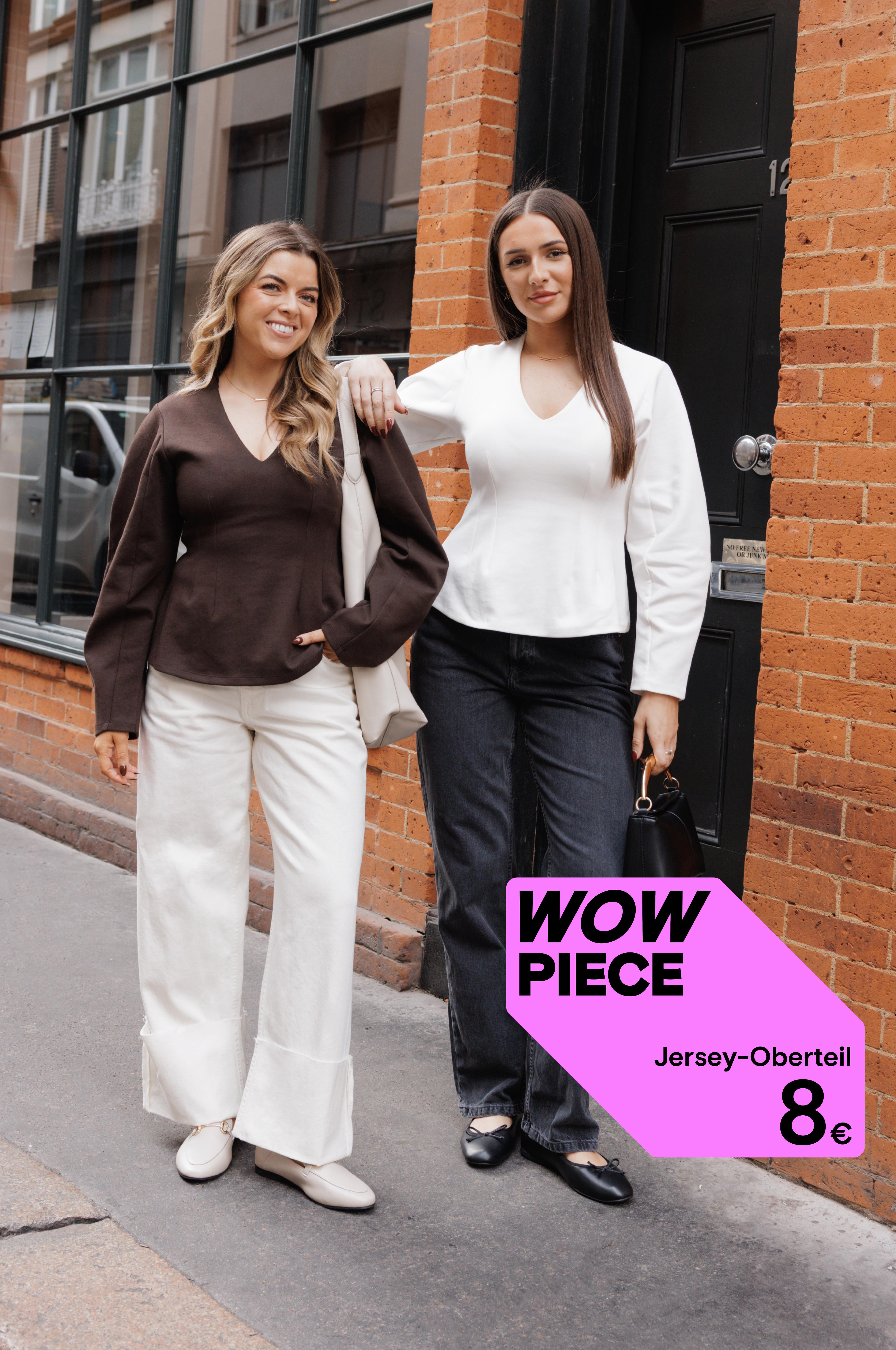 Zwei Frauen in Oberteilen mit V-Ausschnitt vor Backsteingebäude. Eines in Braun mit weißer Hose, das andere in Weiß mit schwarzer Hose.