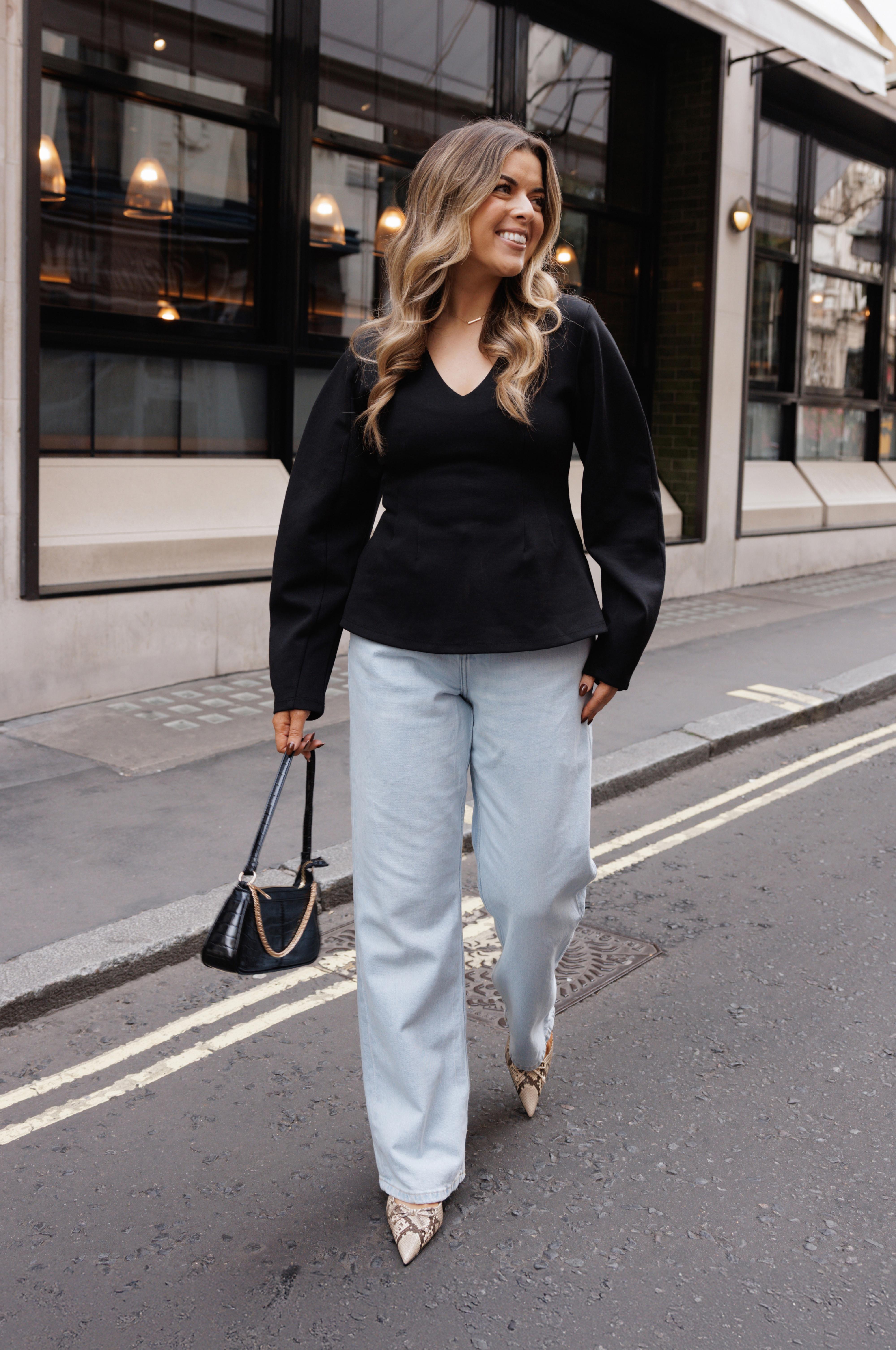 Schwarze Bluse mit V-Ausschnitt und weiten Ärmeln, dazu helle Jeans mit weitem Bein und Heels in Schlangenleder-Optik. Inklusive kleiner schwarzer Handtasche.