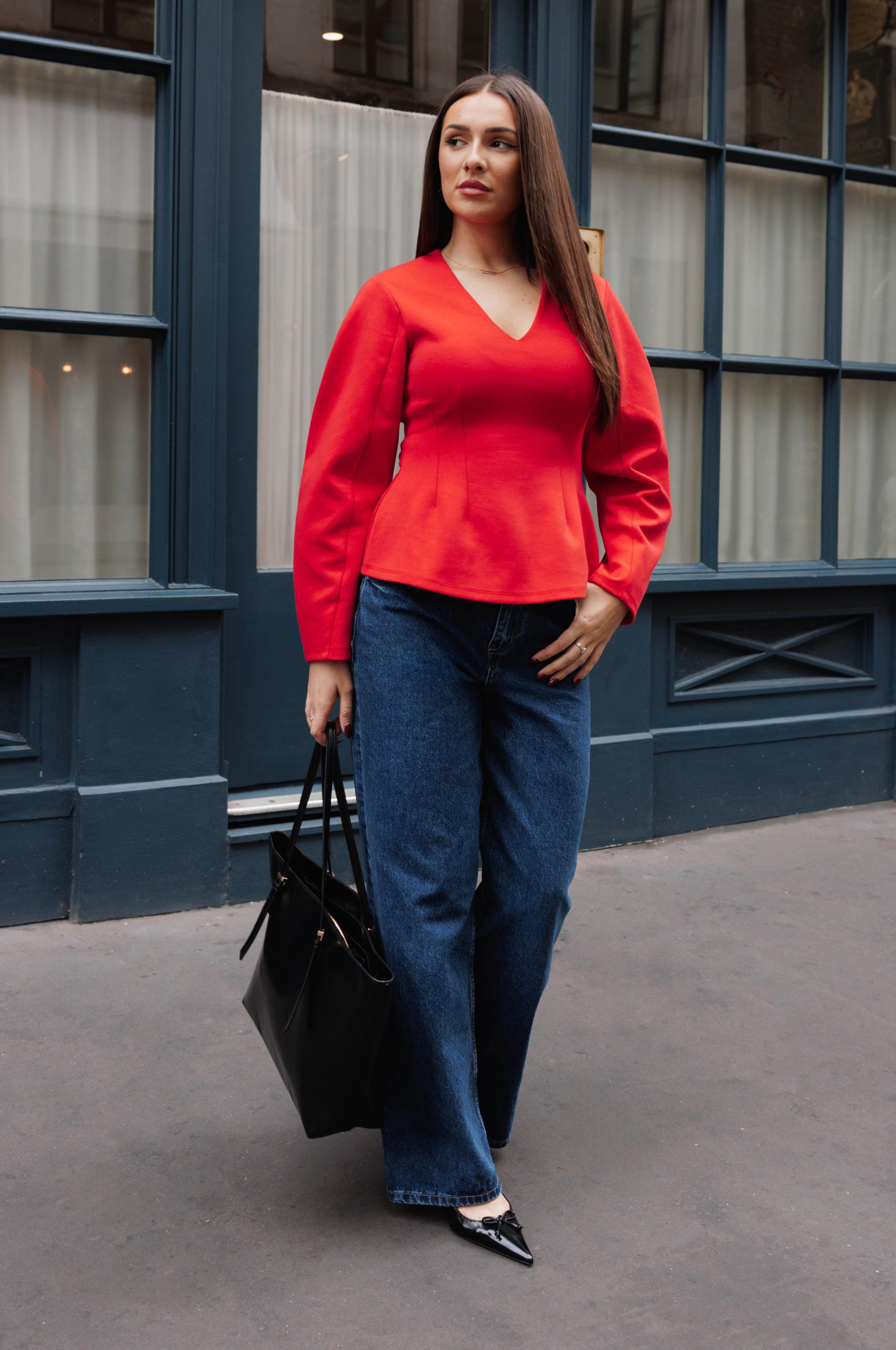 Person in knallrotem Oberteil mit Schößchen und V-Ausschnitt, dazu blaue Jeans und spitze schwarze Schuhe, mit schwarzer Handtasche.
