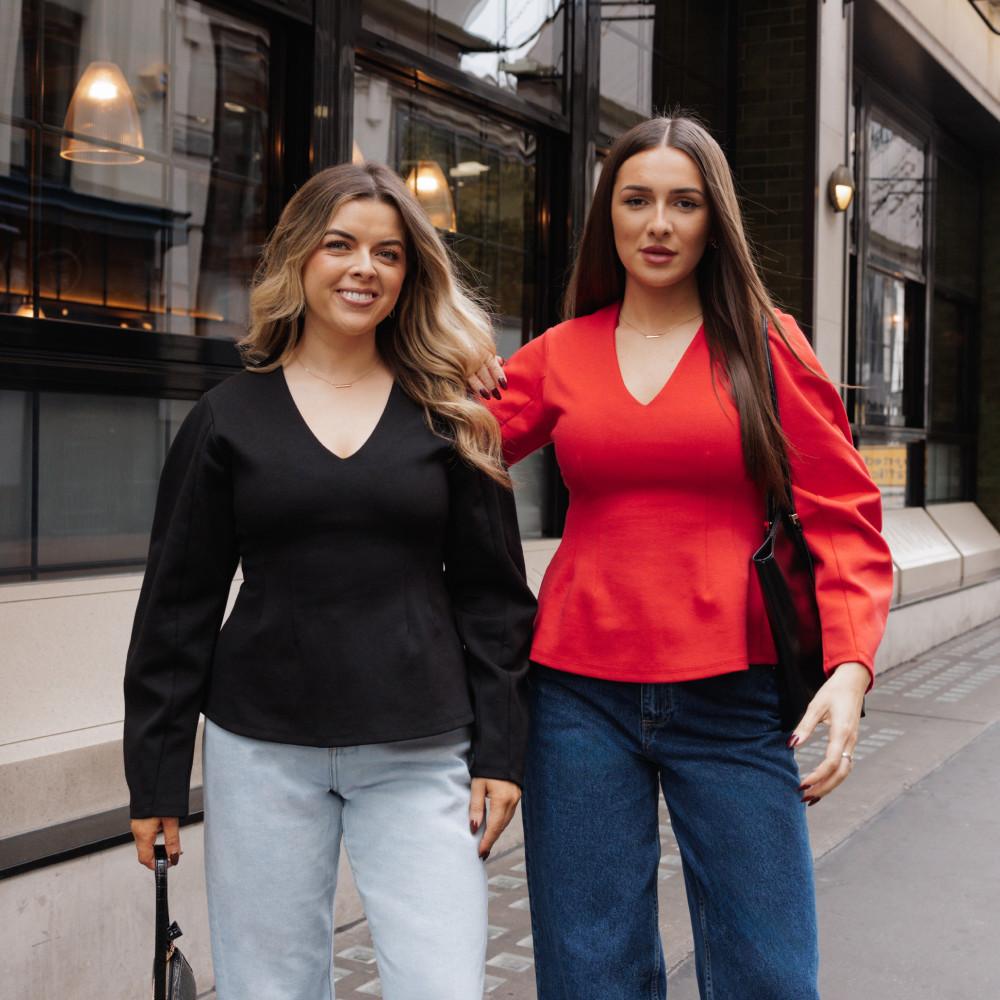 Zwei Frauen in Oberteilen mit V-Ausschnitt, schwarz und rot, kombiniert mit hellen und dunklen Jeans vor einem Café.