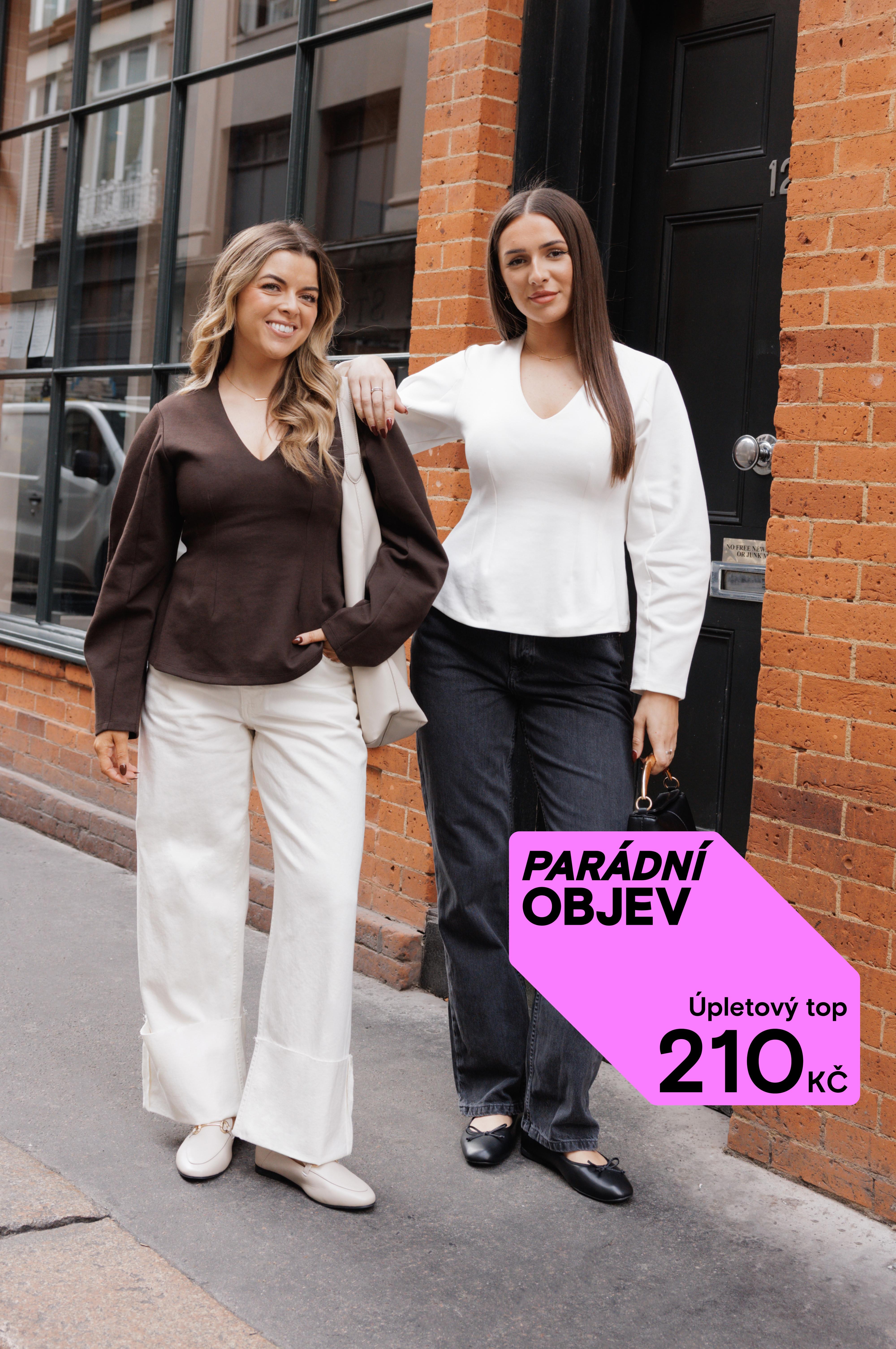 Dvě ženy v topech s výstřihem do V stojí před cihlovou budovou. Jedna má na sobě hnědý top a bílé kalhoty, druhá bílý top a černé kalhoty.